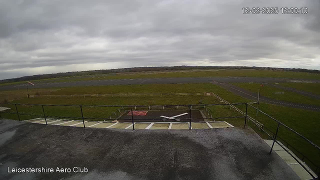 A view of an open grass airfield from a raised platform. The sky is overcast and gray, indicating cloudy weather. In the foreground, there is a flat surface with a white X marking, likely a landing or take-off area. A fence runs along the perimeter of the airfield, and a flagpole is visible on the left side, with a flag fluttering in the wind. The airfield stretches out into the distance, featuring a mix of grassy areas and paved runways.