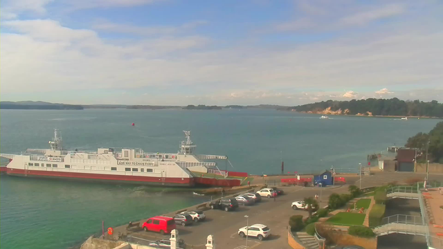 A ferry is docked at a harbor, with its bow facing toward the calm water. The sea is a shade of blue, reflecting the partly cloudy sky. In the foreground, there is a parking lot filled with cars, some white, red, and gray. On the right side, a path leads down to a grassy area with neatly arranged shrubbery and benches. In the distance, a line of trees and a rocky shoreline can be seen, with small boats navigating the water.