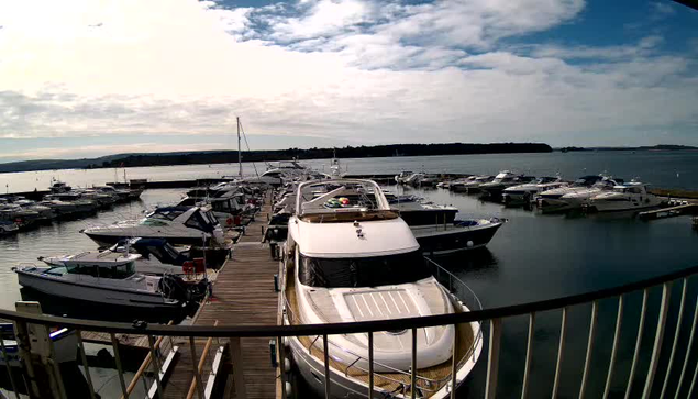 A view of a marina filled with various boats and yachts docked along wooden piers. In the foreground, a large white yacht is prominently positioned. The water is calm and reflects the cloudy sky, which displays hints of blue among the gray clouds. Several smaller boats are visible in the background, and the shoreline features gentle hills. The scene conveys a serene seaside atmosphere.