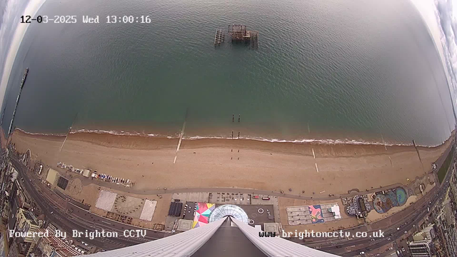 A view from above shows a sandy beach with gentle waves lapping at the shore. The ocean is calm, with varying shades of blue and green. A pier extends into the water on the left side, while small structures are visible in the water slightly further out. The beach area is lined with people and decorative patterns on the sand. Below, there are colorful beach amenities and attractions, including a circular pattern created with sand, along with parked beach chairs. The scene captures a peaceful coastal setting with an overcast sky.