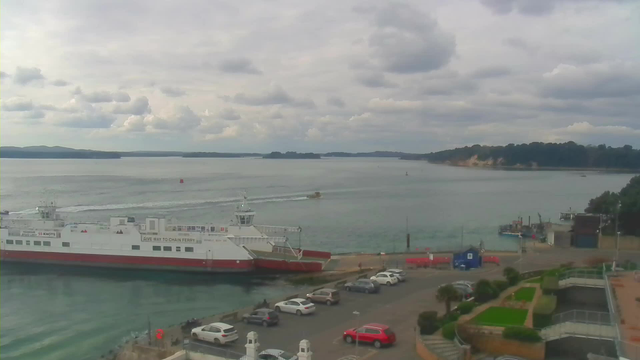 A view of a waterfront scene captured from a webcam. In the foreground, there is a large white and red ferry docked near a parking lot filled with various cars. The parking area features a small garden with green plants. Beyond the ferry, a calm body of water stretches out, with several small boats and distant islands visible. The sky is partly cloudy, with soft, gray clouds. The outline of a shoreline with lush greenery is visible in the background.