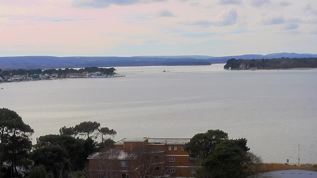 A scenic view of a calm body of water, with a shoreline featuring trees and buildings in the foreground. In the distance, several boats can be seen on the water, with hills and a cloudy sky providing a backdrop. The overall atmosphere is serene, with gentle waves reflecting the muted colors of the sky.