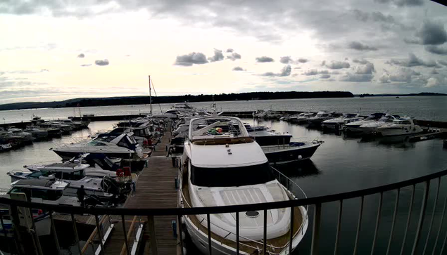 The image shows a marina filled with various boats and yachts docked along a wooden pier. In the foreground, a large white yacht with a light-colored deck is prominent. The water is calm, reflecting the cloudy sky above, which features a mix of gray and white clouds. Several smaller boats are visible in the background, and the shoreline can be seen in the distance, lined with trees. The overall atmosphere is serene, with a hint of overcast weather.