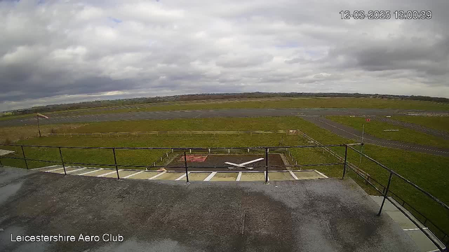 A cloudy sky is overhead, with gray clouds covering most of the view. Below, there is an airfield with a patch of green grass prominently featured. To the left, a small section of the runway is visible, along with a red and white windsock on a pole. In the foreground, there is a concrete deck with a white "X" marking on it, indicating a landing area. On the lower left, the text "Leicestershire Aero Club" is visible. An empty, open space stretches out in front of the deck, leading to the runway in the distance.