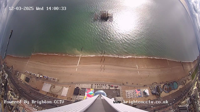 Aerial view of a beach with golden sand and clear greenish water. In the distance, a pier extends into the ocean, while a partially submerged structure is visible just offshore. The beach is lined with people walking along the shore. Below, various amusement park rides and structures can be seen, along with colorful canopies. The sky is mostly clear, and the scene is illuminated by daylight. A timestamp in the corner indicates the date and time.