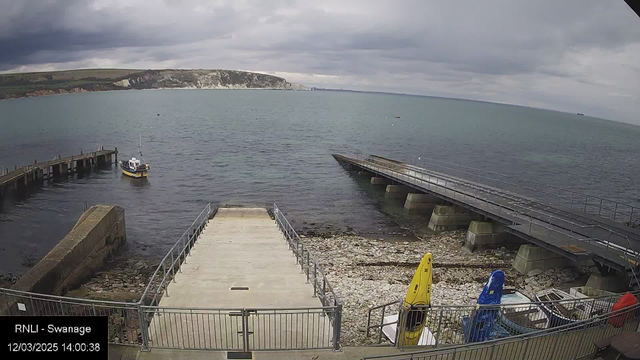 A view of a body of water with a small boat near a pier. To the left, there is a rocky shoreline and a concrete ramp leading down to the water. In the foreground, several kayaks in bright colors, including yellow and blue, are stored near the ramp. The background features a cloudy sky with grey clouds, and a distant coastline with cliffs is visible across the water. The scene appears calm with gentle ripples on the surface of the water.