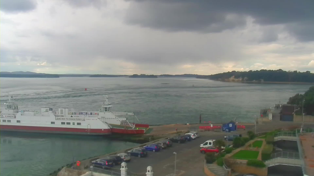 A cloudy sky hangs over a body of water where a large ferry is docked at a terminal. The ferry has a white body with a red stripe, and a few people can be seen on board. To the right, there is a parking area filled with various vehicles. In the background, green hills can be seen along the shoreline, with trees lining the water's edge. The water is rippling, possibly due to currents, and small buoys are visible in the distance.