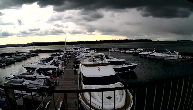 A marina filled with various boats is seen from above, with a wooden dock extending into the water. The sky is overcast with dark clouds, and a sunset casts a subtle glow on the horizon. Several boats are moored in the water, some of which are larger yachts, while others are smaller vessels. The water appears calm, reflecting the cloudy sky.