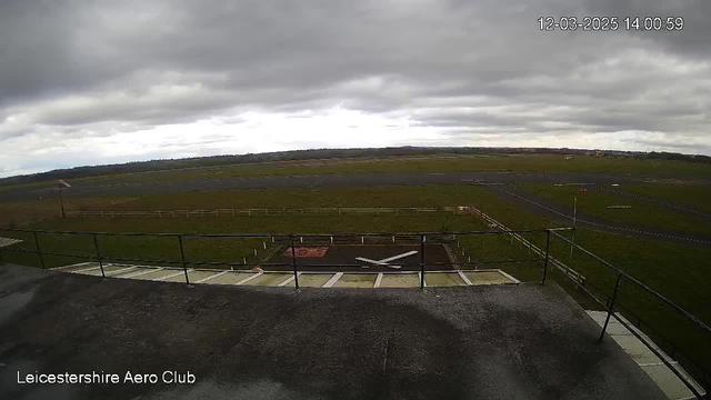 A view from a webcam at Leicestershire Aero Club showing a large outdoor space. In the foreground, there is a flat surface with a visible cross marking, along with other ground markings. Beyond that, an open field extends, leading to a runway with a few planes parked in the distance. The sky is mostly cloudy, and there are scattered patches of grass surrounding the area. The image reflects a dull, overcast day.