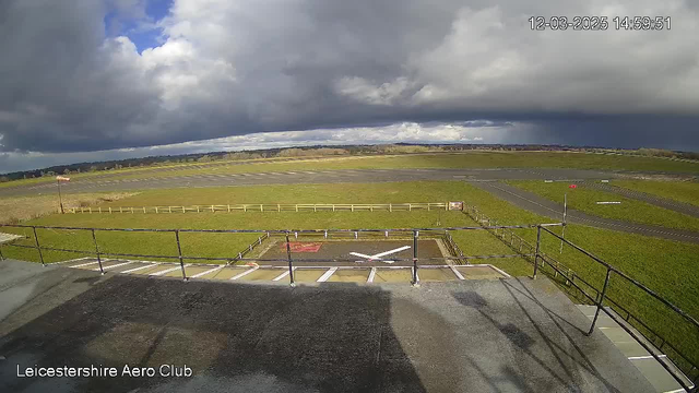 A view from a webcam at Leicestershire Aero Club showing an airfield under a partly cloudy sky. The foreground features a grassy area with a light-colored fence and a marked landing zone with a red and white cross. In the background, there are runways with a few markings and some distant hills under a blue and gray sky.