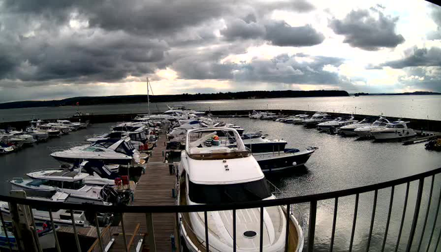 A marina scene with multiple boats docked in calm water. The sky is overcast with dark clouds, creating a moody atmosphere. In the foreground, a large white boat is prominent, with smaller boats visible around it. The marina is lined with a wooden walkway, and distant hills are seen on the horizon.