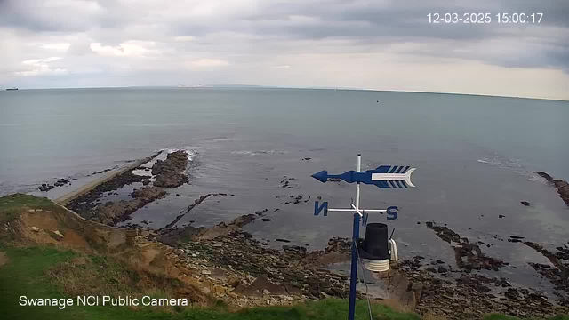 A coastal scene shows a rocky shoreline extending into a calm, flat sea under a cloudy sky. In the foreground, there is a blue and white weather vane with directional indicators pointing north, south, and west. The camera is positioned at a higher vantage point, providing a panoramic view of the water and land. The image includes a timestamp at the top right indicating the date and time.