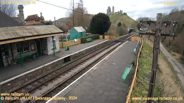 A view of Corfe Castle railway station on a cloudy day. The platform features a waiting area with green benches, a stone wall, and a wooden fence. In the background, a hill rises with the ruins of Corfe Castle visible atop it. There is a small green shed and a sign indicating the way out. Train tracks are visible in the foreground, with gravel and concrete surrounding them. A utility pole stands on the right side of the image, with wires extending above.