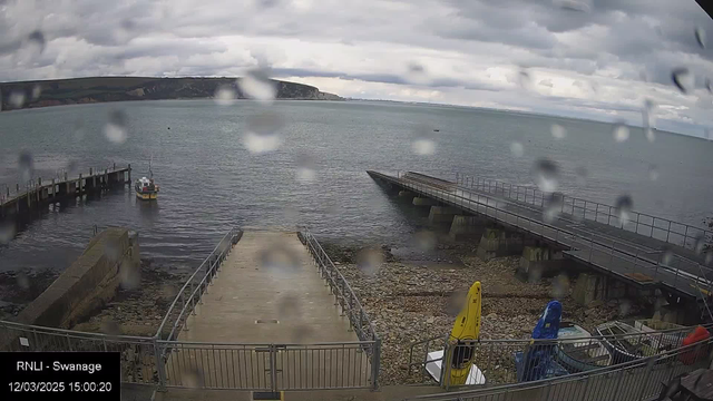 A foggy view of a marina by the sea. In the foreground, there are two ramps leading down to the water, with a small fishing vessel docked near the left side. The surface of the water is calm but rippling, reflecting gray clouds above. Rains droplets are visible on the camera lens, creating a hazy effect. On the right side, there are several kayak-like boats stored upright, colored yellow and blue. The distant shoreline is lined with green hills under the cloudy sky.