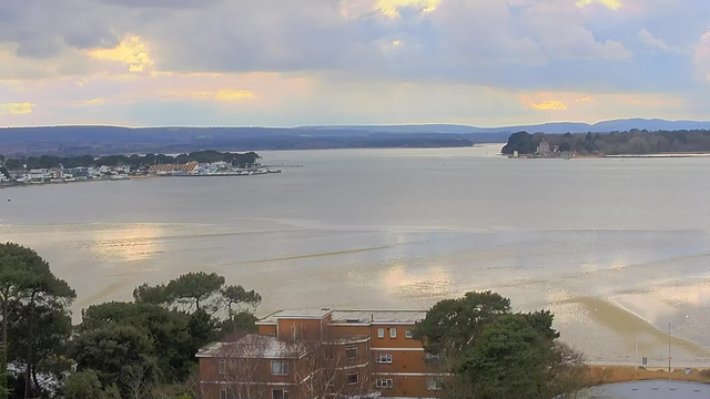 A panoramic view of a calm body of water under a cloudy sky. In the foreground, there are trees and a building with a flat roof. The water reflects soft colors from the sky, with gentle ripples visible. In the background, a small town with white buildings and some boats can be seen along the shoreline. Further away, hills rise in the distance, under a mix of gray and light blue clouds. The overall atmosphere is serene and tranquil.