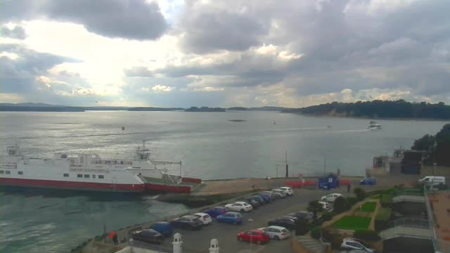 A view of a calm waterway with varying shades of grey clouds above. On the left, a ferry with red and white colors is docked. There are several boats in the water, including a larger yacht moving towards the right. A parking lot with numerous cars in different colors is visible in the foreground. To the right, a small pier can be seen, and there are green shrubs and landscaping elements nearby. The distant shoreline features trees and hills under the cloudy sky.