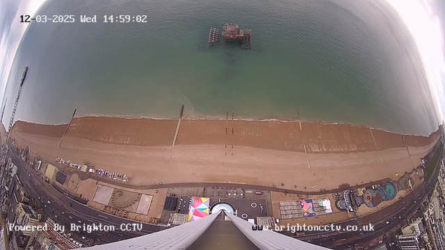 A high-angle view of a beach scene with a sandy shoreline and shallow water. In the background, a pier extends into the sea, partially submerged. The beach is mostly empty, with a few people walking along the shore. Below, an amusement park with colorful structures and pathways is visible. The time and date are displayed at the top of the image. The overall atmosphere is calm, with clouds in the sky above.