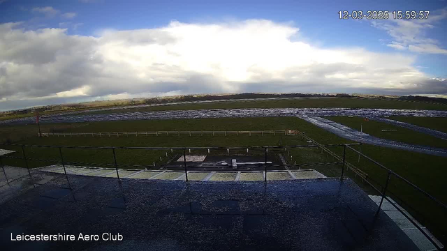 A wide view from a webcam showing an airfield under a cloudy sky. The foreground features a railing, leading to an open grassy area with some wet patches, indicating recent rain. In the distance, a long stretch of asphalt runway is visible, bordered by a white fence. The time and date are displayed in the corner of the image.