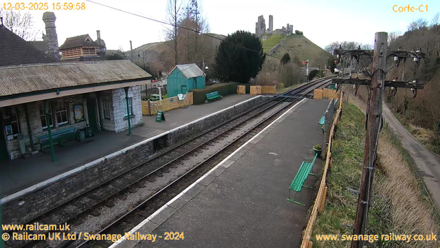 A view of a railway station in a rural setting. The foreground shows a platform with a few green benches and a sign that says "WAY OUT." To the left, there is a stone building with large windows, indicating the station house. In the background, a hill rises, topped with the ruins of a castle. The area around the station is surrounded by trees and shrubs, with additional seating near a wooden fence. The railway tracks run parallel to the platform, extending into the distance. The sky above is clear with a hint of clouds.