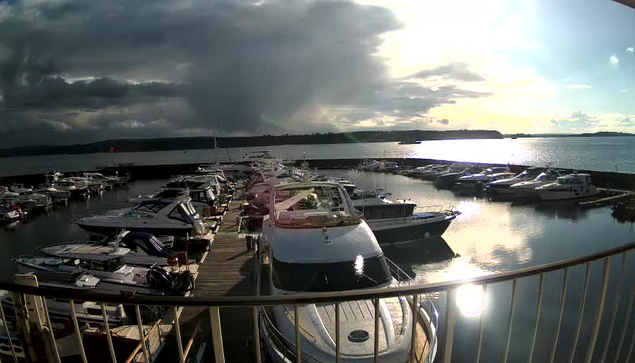 A marina scene with several boats docked at a pier. The water is calm and reflects the sky, which is partially cloudy with a bright sun. The shoreline is visible in the background, with trees and hills. Some large yachts are in the foreground, along with smaller boats. The overall atmosphere is tranquil, with gentle waves lapping at the dock.