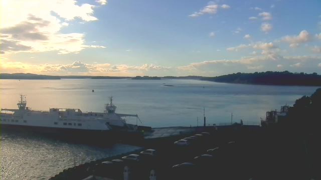 A serene view of a harbor at sunset, featuring a large white ferry docked at the quay. The water is calm and reflects the colorful sky, which includes soft clouds illuminated by the setting sun. In the distance, islands can be seen along the horizon, and several parked cars line the edge of the harbor. The atmosphere is peaceful, with gentle waves and a clear blue sky transitioning to warm hues.