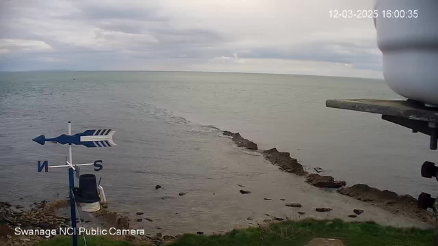 A coastal scene showing calm water extending to the horizon under a cloudy sky. In the foreground, there's a weather vane with a blue arrow pointing north, and a small rocky shoreline is visible on the right. The camera is mounted on a structure, with a white container partially visible at the top. The timestamp indicates the image was captured on March 12, 2025, at 4:00 PM.