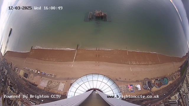 A high-angle view of a sandy beach and the ocean. The shoreline is visible, with a pier extending into the water. Below, a circular structure appears at the edge of the image, possibly a viewing platform. The beach has people in the distance, and colorful buildings and amusement rides can be seen along the beachfront. The ocean is calm, blending from light blue to deeper green shades.