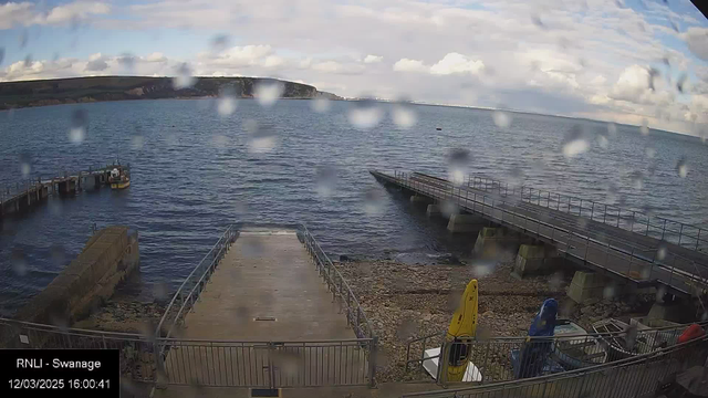 A waterfront scene is shown with several piers extending into a calm body of water. In the foreground, a wide ramp leads down from the viewing area to the water's edge, with rocky shoreline visible beneath it. To the right, a few colorful kayaks are stored upright on the ground, with yellow, blue, and red colors visible. In the background, the water is calm and reflects a partly cloudy sky, with some raindrops visible on the camera lens, slightly obscuring the view. A small boat is docked at the end of one of the piers, and distant cliffs can be seen along the horizon.