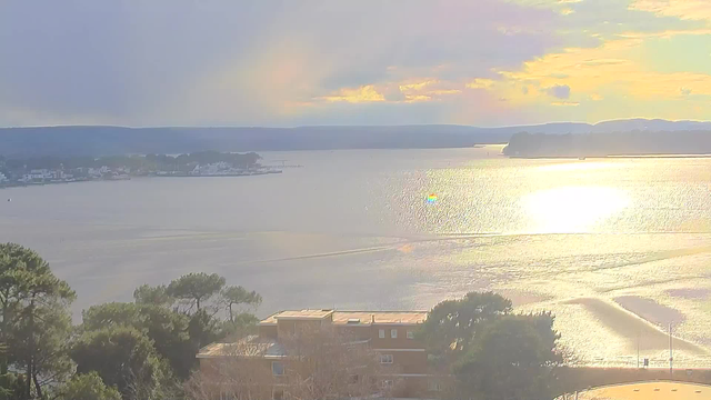 A serene landscape featuring a body of water reflecting sunlight, surrounded by distant hills. In the foreground, there are trees and buildings, with a small coastal town visible on the left side. The sky displays soft clouds with shades of blue and yellow. The overall scene conveys a tranquil, natural atmosphere.