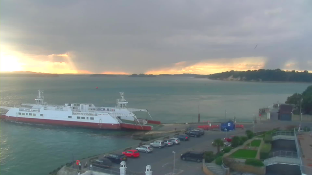 A ferry docked at a pier in the foreground, with various parked cars nearby. In the background, the water reflects a fading sunset with clouds above, while distant land outlines the horizon. The scene has a calm atmosphere with soft light breaking through the clouds.