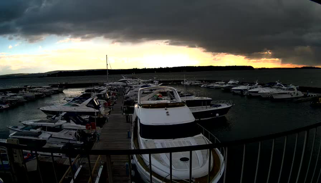 A marina filled with various boats docked at a wooden pier. In the foreground, a large white boat is prominently positioned, its shiny surface reflecting the dim light. Surrounding boats include smaller vessels of different shapes and colors, mostly white and blue. The sky above is dark and cloudy, with shades of gray and hints of orange as the sun sets in the background. The water is still, with a smooth surface mirroring the colorful sky.