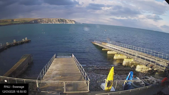 A view of a coastal scene featuring a calm blue sea under a partly cloudy sky. The left side shows two wooden piers extending into the water. In the foreground, two colorful kayaks—one yellow and one blue—are secured by the shore, while a rocky beach is visible nearby. The distant coastline has grassy hills leading up to white chalk cliffs, contrasting with the darker water. The image is timestamped with the date and time at the bottom left corner.