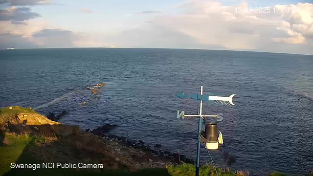 A view of the sea from a webcam, showing calm waters with gentle waves. In the foreground, there is a grassy area alongside rocky shoreline. A weather vane is positioned to the right, featuring a blue arrow pointing north and a design of a fish on the horizontal arm. The sky is partly cloudy with patches of blue and sunlight illuminating the scene. There are distant boats visible on the horizon. The timestamp at the top right indicates the date and time.