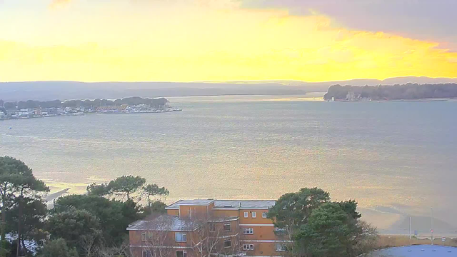 A tranquil view of a body of water during sunset, with golden and orange hues reflecting on the surface. In the foreground, there are several trees and a brown building. Across the water, a shoreline is visible with various structures, including a marina with boats. The background features a low range of hills under a cloudy sky.