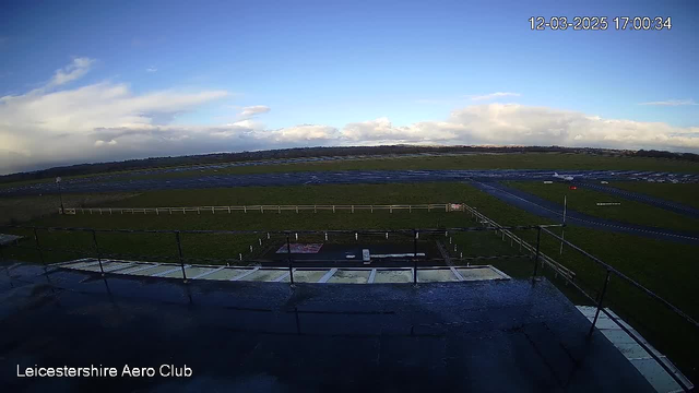 A clear sky with sparse clouds is visible above a wide open field, which is part of an aerodrome. The ground is predominantly green grass, with a dark asphalt runway running through the center. In the foreground, there is a railing along the edge of a building, hinting at an observation deck. Several white poles are scattered along the perimeter of the field. Overall, the scene is bright and well-lit, depicting a tranquil outdoor setting.