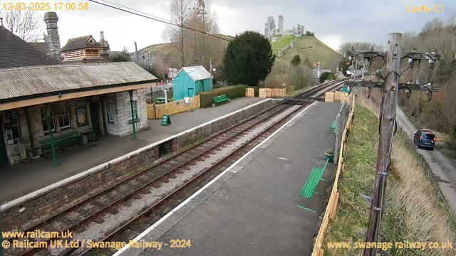 A train station scene shows a platform with two sets of railway tracks. On the left, there is a stone building with a green bench and a poster on the wall. Several green benches are placed on the platform. In the background, there is a hill with ruins of a castle at the top. A blue-green wooden building is visible on the platform, and a sign that reads "WAY OUT" is positioned on the ground. On the right, there is a wooden fence and a road where a car is visible. The sky is cloudy.