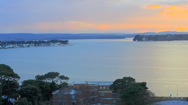 A panoramic view of a calm body of water during sunset, with gentle waves reflecting soft hues of orange and pink in the sky. In the foreground, there are dark green trees and buildings, while the shoreline features a town with residential and commercial structures. The horizon is lined with rolling hills, and small boats can be seen near the shore, adding to the tranquil atmosphere.