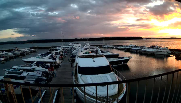 A marina at sunset, featuring numerous boats docked in calm water. The sky is filled with dramatic clouds blended with shades of orange, pink, and purple as the sun sets on the horizon. A wooden dock is visible in the foreground, along with a white yacht that appears prominently from the left side of the image. The overall atmosphere conveys a peaceful waterfront scene.