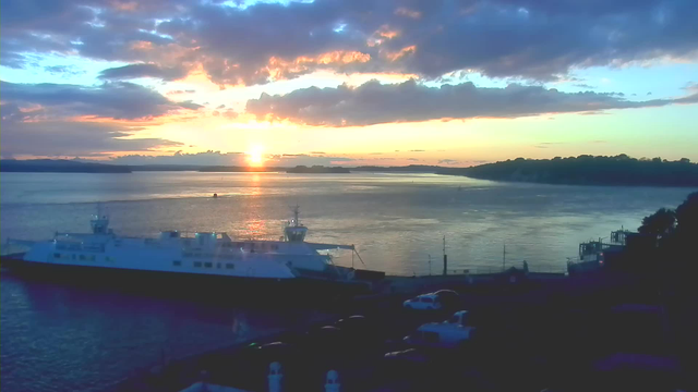 A scenic view of a harbor at sunset. The sun is setting near the horizon, casting a warm golden light over the water, which reflects the colors of the sky. There are several clouds in various shades of blue and orange. In the foreground, a large ferry boat is docked, and there are a few vehicles parked near the water. The landscape in the distance features tree-covered hills silhouetted against the fading light of the sky.