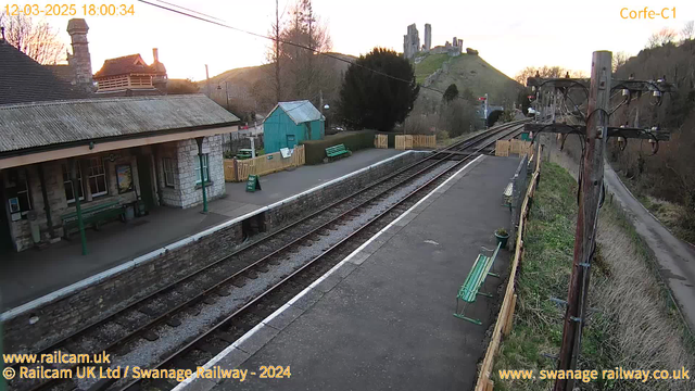 A train station platform is depicted, with two sets of railway tracks running parallel on the right side. The platform features several green benches and a building with a stone facade and a sloped roof in the foreground. A green shed is visible in the middle ground, accompanied by a wooden fence and shrubs. In the background, there are hills with ruins or structures atop them, illuminated by a setting sun. The sky shows hues of orange and pink, indicating twilight. A wooden utility pole stands on the right side, with visible wires extending overhead.