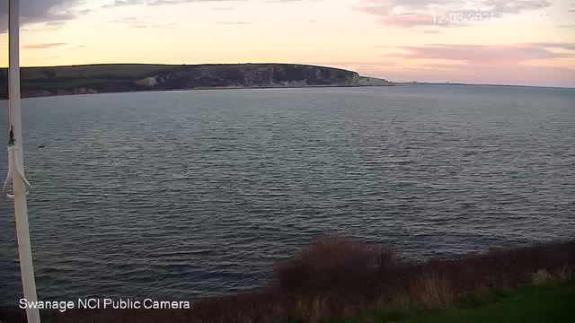 A coastal scene captured from a webcam shows a body of water extending towards the horizon. Gentle waves reflect the soft colors of the sky above, which is transitioning from daylight to twilight with hues of pink and orange. In the distance, a rocky cliff rises along the shoreline. The foreground features some grass or low vegetation. The image is serene, indicating a calm atmosphere.