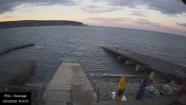 A view of a harbor at dusk. In the foreground, there is a flat, concrete ramp leading down to the water, surrounded by railings. To the right, a wooden pier extends into the water, with sections visible above the surface. On the left side, rocky shoreline is visible, with several brightly colored kayaks (yellow, blue, and red) positioned beside the ramp. The water is calm, reflecting the colors of the sky, which transitions from soft pink to gray as the sun sets in the background. A distant cliff and landmass can be seen on the horizon, under a partly cloudy sky.