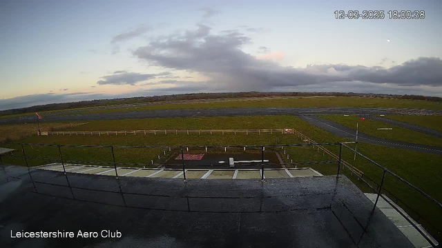 A view from a webcam showing the Leicestershire Aero Club. The scene features a large grassy area with a paved runway visible. There are clouds in the sky, creating a mix of light and shadow across the landscape. A small fence is seen surrounding the grassy area, and a few objects are visible on the ground near the center, including a rectangular red surface. The sky is lightening toward the horizon, indicating a time near sunset.