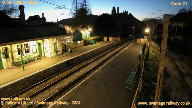 A railway station in the early evening, featuring a stone building with large windows and a green door on the left. There are green benches along the platform. The train tracks run diagonally through the image, with a lamppost illuminating the area. In the background, a hill features a silhouette of castle ruins against a twilight sky. A sign that reads "WAY OUT" is positioned near the entrance to the station. The scene is mostly calm, with no trains present.