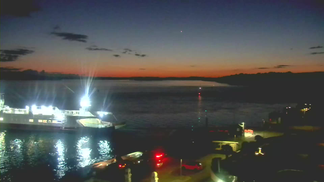 A twilight scene featuring a calm body of water reflecting soft light. A large ferry with bright lights is docked on the left side, surrounded by dark water. The horizon displays a gradient of colors, transitioning from deep blue to warm orange and soft pink. Small lights indicate activity at the dock, and a few ruffled clouds are visible in the sky.