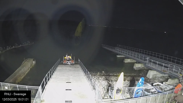 A nighttime scene at a dock, showing a rescue boat with two people in yellow vests on a ramp leading toward the water. The ramp is surrounded by a metal railing. To the right, there are several colorful kayaks on the shore, and a calm body of water is visible. The sky is dark, and the image shows limited visibility, with faint lights in the distance across the water.