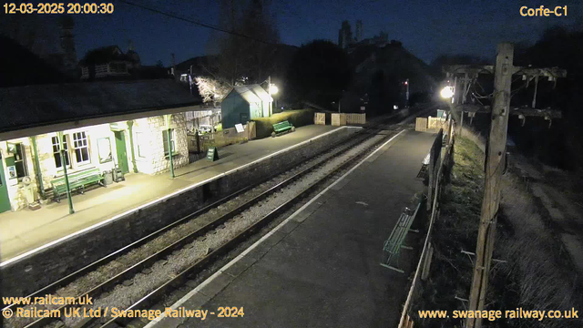 The image shows a train station platform at night. To the left, there are two stone buildings with green trim, illuminated by warm lights. Benches are visible on the platform, and a sign reads "WAY OUT." The nearby surroundings include grass and trees. On the right, there is a wooden fence, and a tall wooden pole with electrical lines runs vertically. The railway tracks stretch into the distance, surrounded by gravel and asphalt. The sky is dark, indicating nighttime.