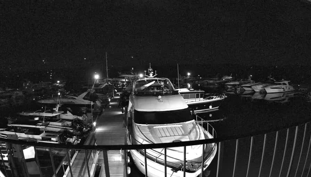 A nighttime marina scene with multiple boats docked in a harbor. The image is primarily in black and white, with some dimly lit areas showing lights reflecting off the water. A large yacht is positioned prominently in the foreground, with various smaller boats visible in the background. The scene conveys a sense of calm and tranquility, typical of a quiet night at the docks.