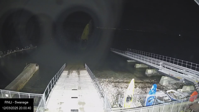 A nighttime view of a harbor area with a wooden ramp leading to the water, surrounded by dark water and rocky shoreline. On the left side, there is a concrete pier extending into the water, with some mooring posts visible. Several small boats, including yellow and blue kayaks, are positioned on the right near the shore. The scene is illuminated by artificial lights, creating a subtle reflection on the water. The sky is dark, indicating it is nighttime.