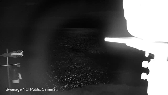 A dark image of the sea at night, with faint reflections of light on the water's surface. In the foreground, there is a weather vane and a pole, suggesting a coastal location. The overall scene is moody, with limited detail visible due to the low light.